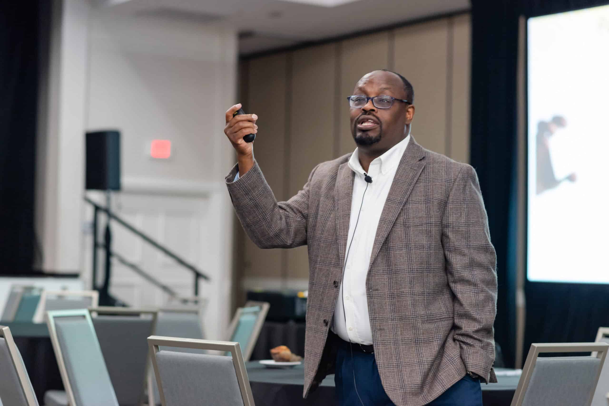 A Black professional man stands at the front of the room with a presentation clicker. He seems to be giving a presentation.